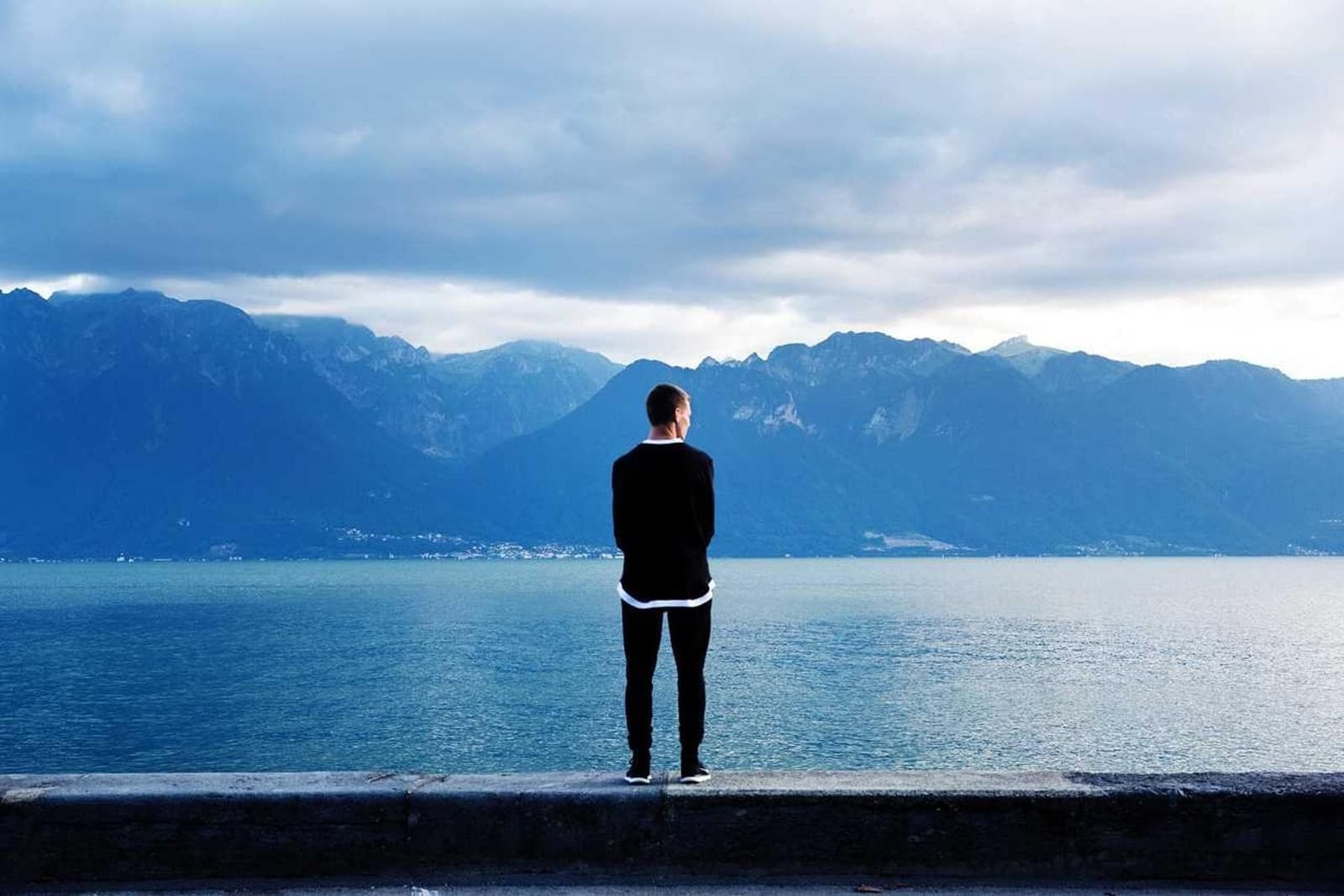 A person standing by a lake below mountains.